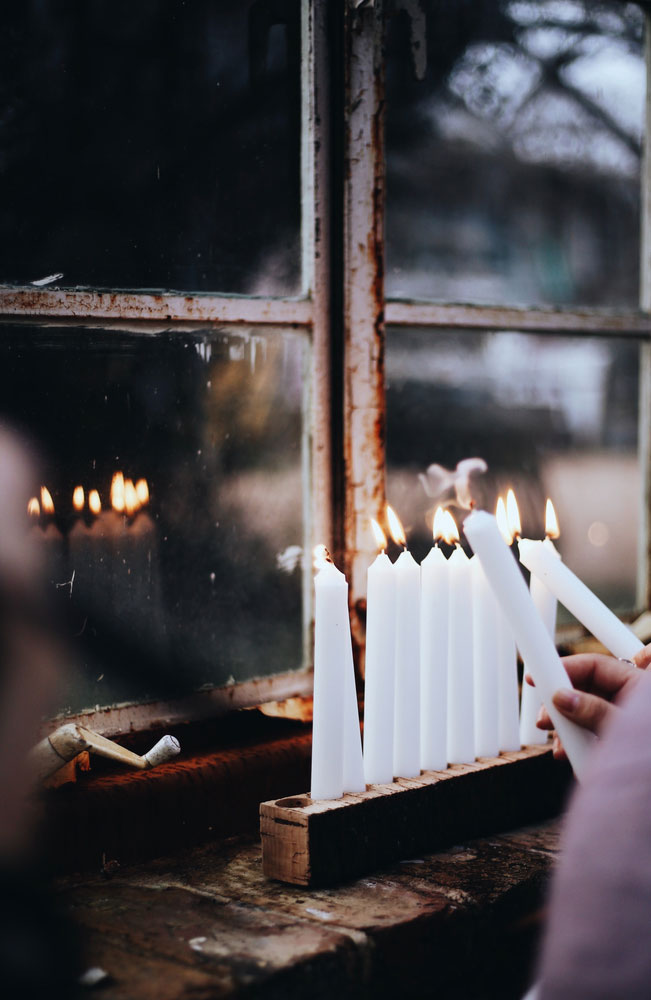 white candles in a row set on a window sill, a person is lighting one of the unlit candles